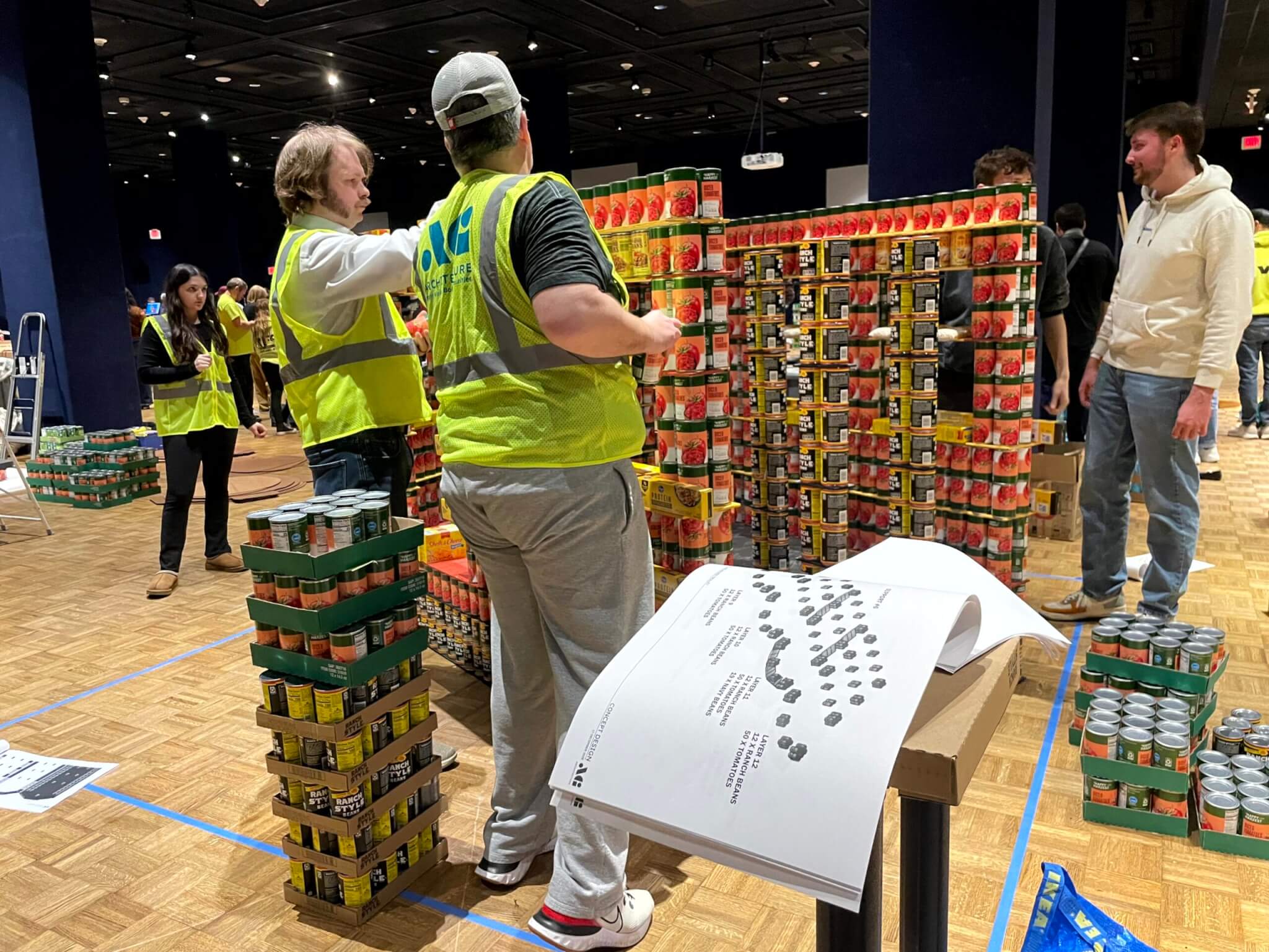 CANstruction building continues with AG's team referencing their printed plans for exact placement of cans and boxes.
