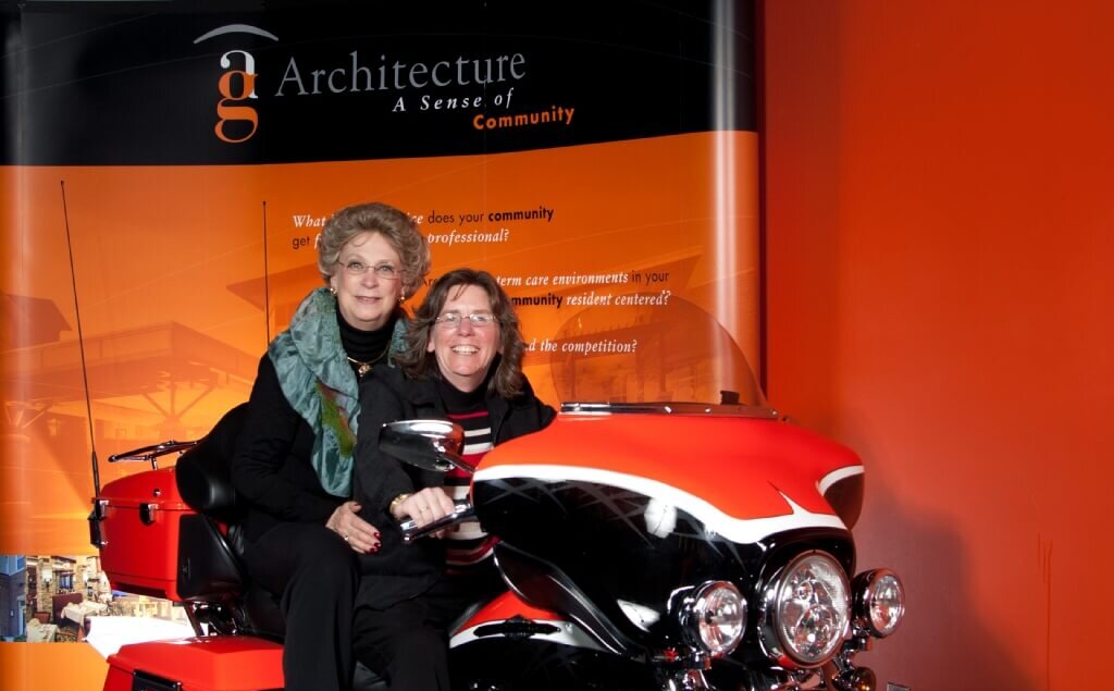 A photo of Cindy Lofy and Carole Hoppe on a motorcycle at the AG 20th Anniversary party at Harley Davidson in 2012.